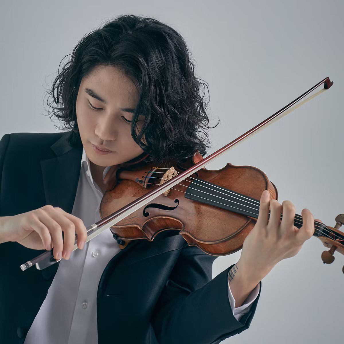 Musician in a suit playing a violin, eyes down and bow in hand against a light gray background.