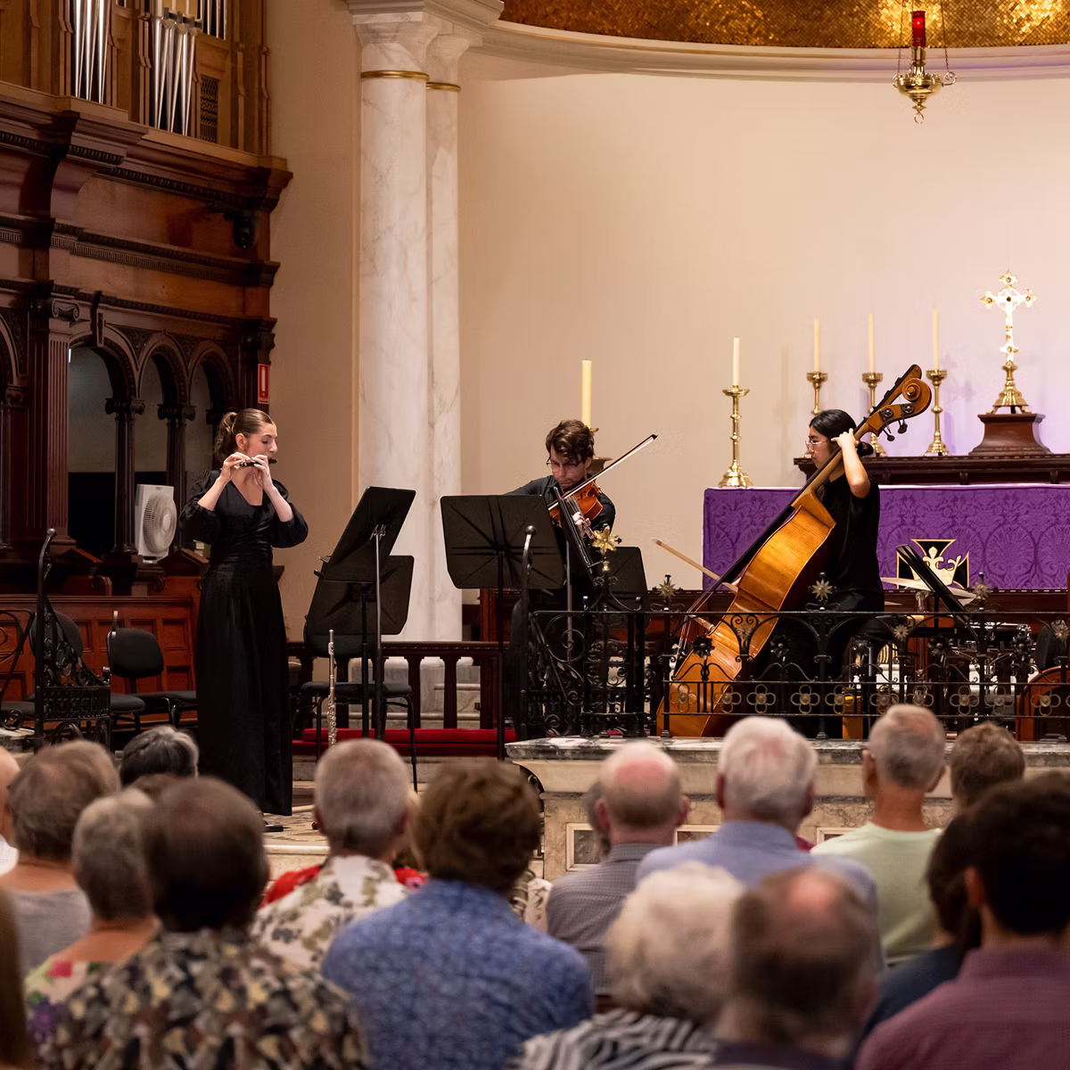 Musicians perform in a church during a concert, candles and purple altar cloth behind seated audience in foreground.