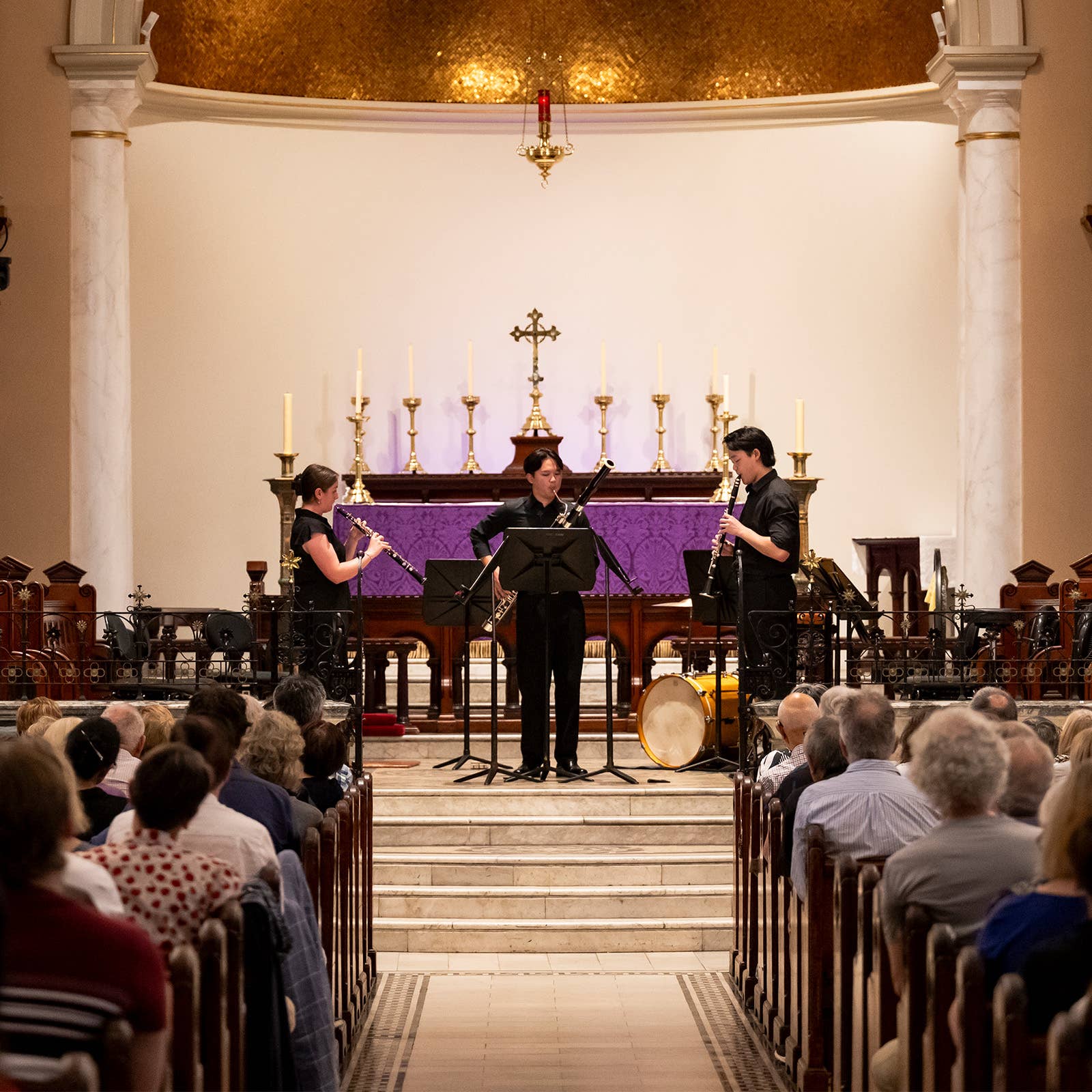Musicians in black play trumpets and percussion on a church stage before a purple altar with candles