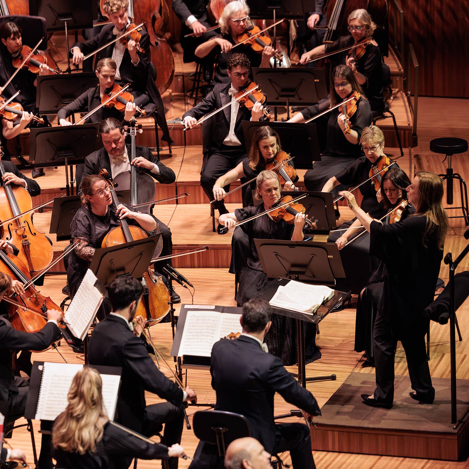 Orchestra members in formal attire playing violins and cellos on stage during a live performance.