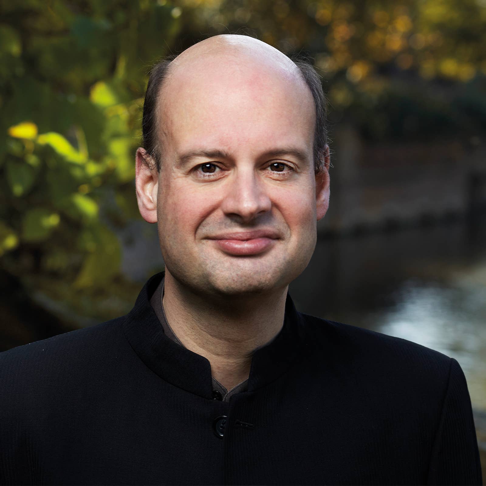 Smiling man with balding hair in a dark jacket against a softly blurred outdoor park background