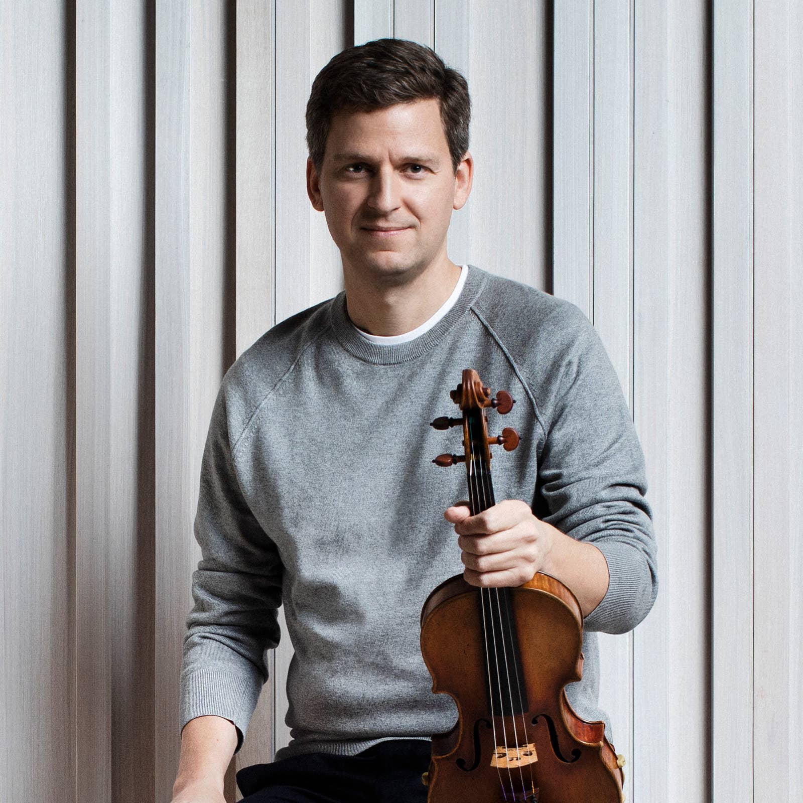 Smiling man holding a wooden violin, standing against light paneled wall.