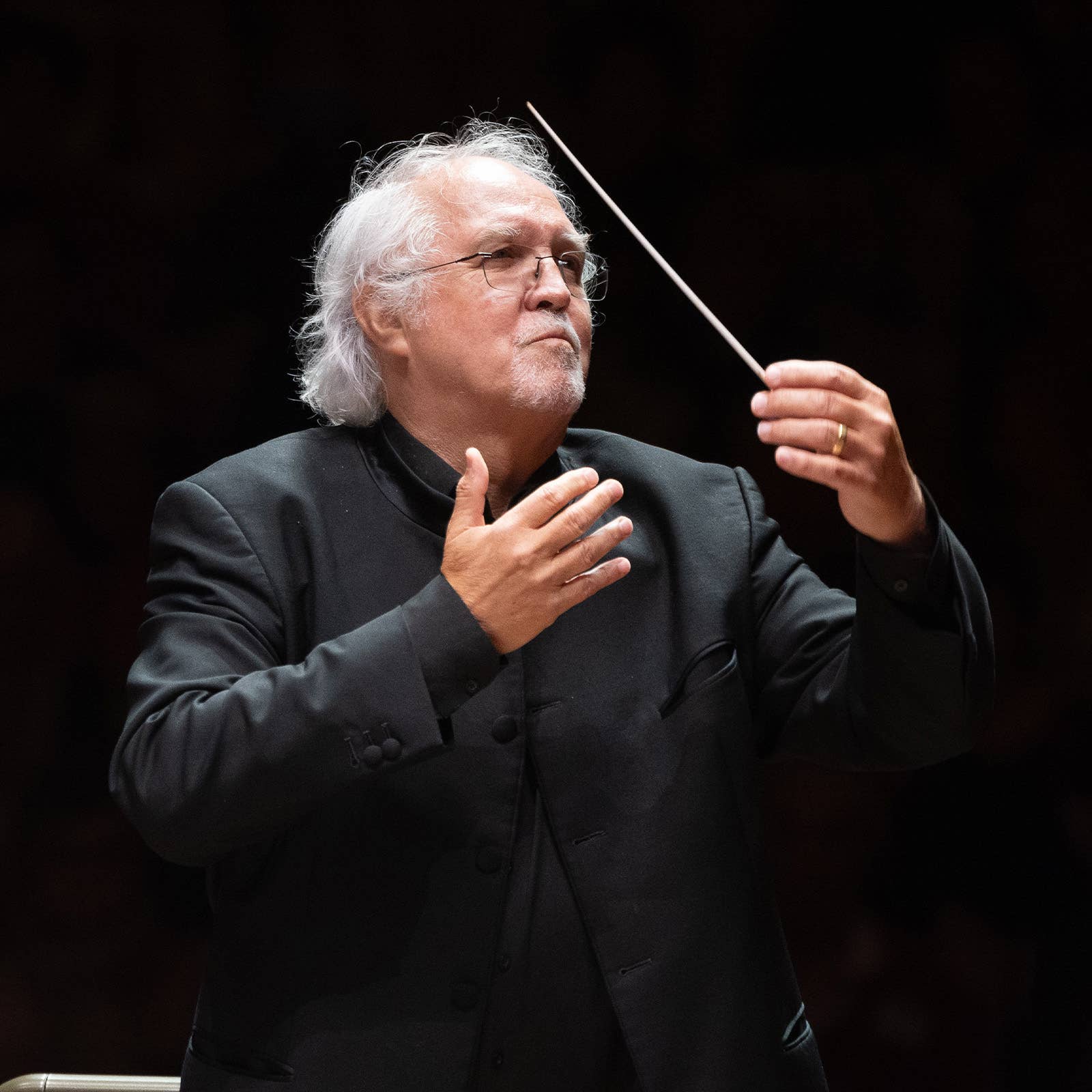 Elderly male conductor with gray hair and glasses, holding a baton and gesturing during a performance.