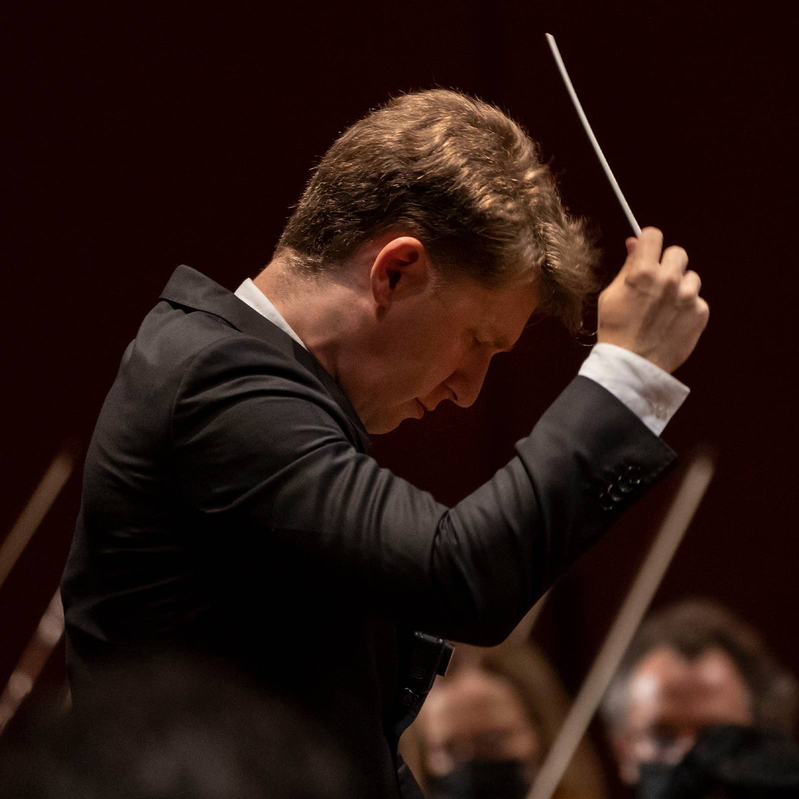 Male conductor in a dark suit holding a baton, directing an orchestra under warm stage lighting.