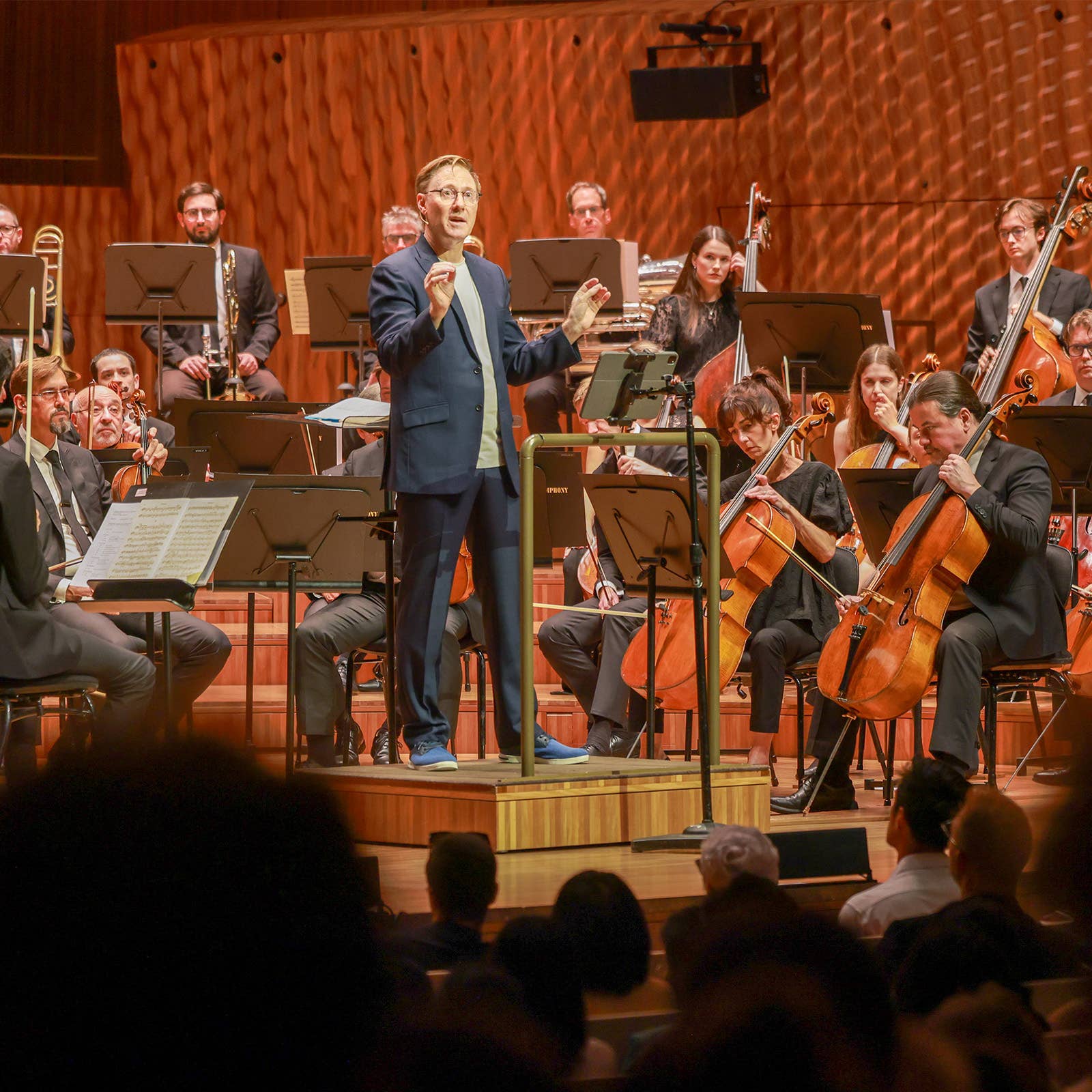 Conductor leads an orchestra onstage with brass, violins, and cellos under warm stage lights.
