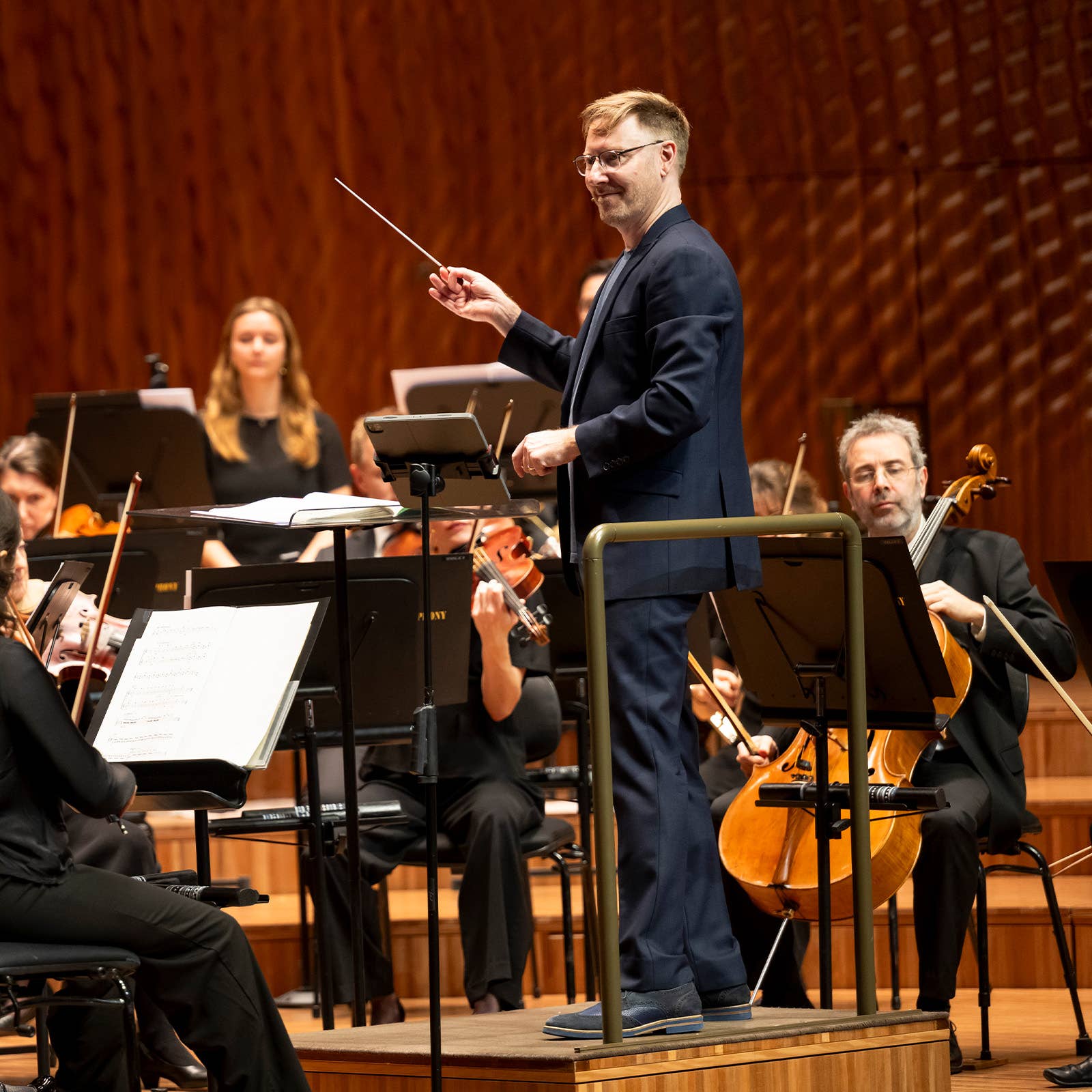 Conductor leads an orchestra on stage, holding a baton while musicians play amid music stands.