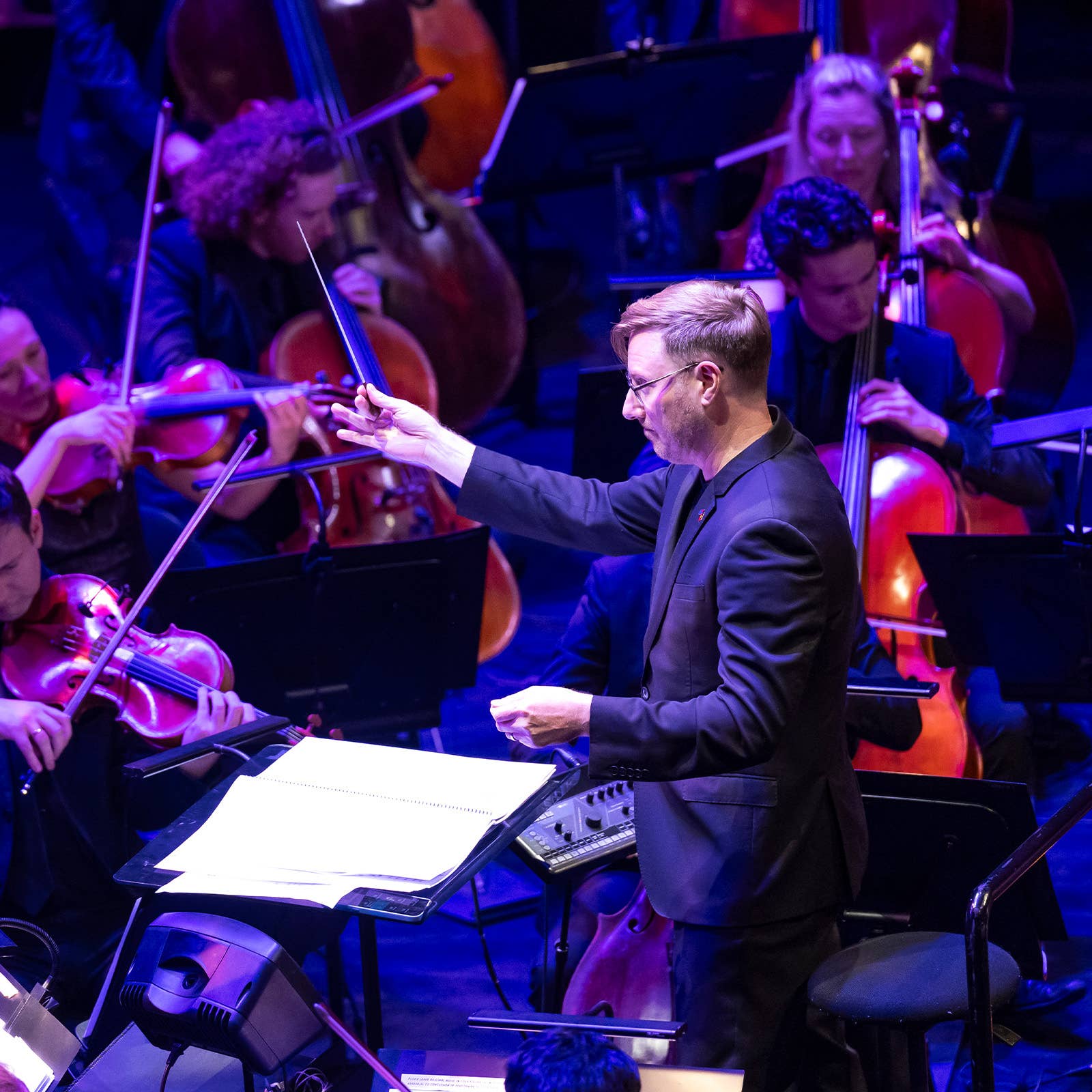 Conductor in suit leads an orchestra as violinists and double bass players perform under blue stage lights
