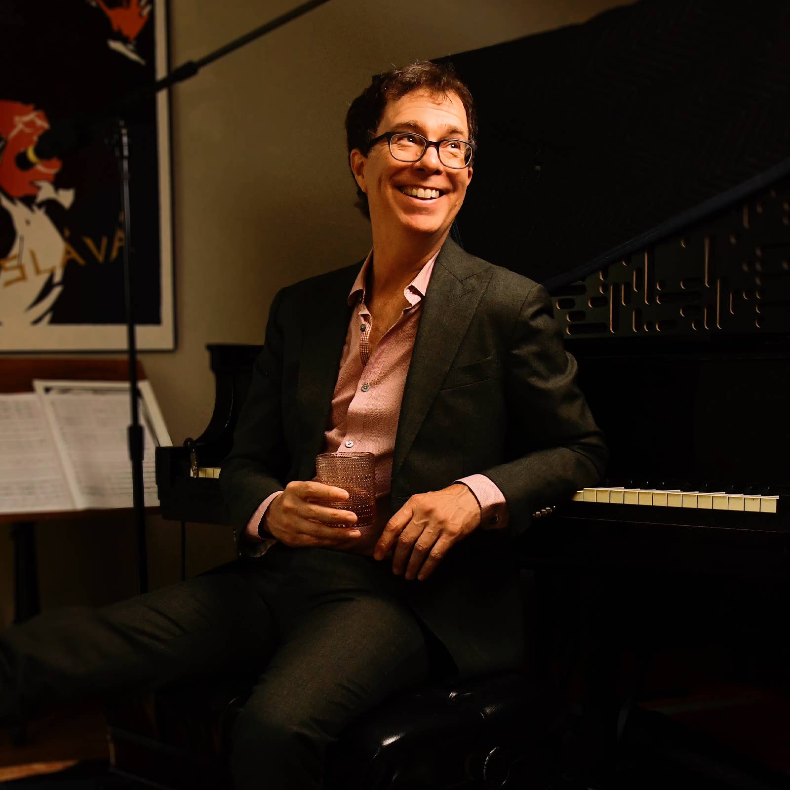 Smiling man with glasses seated beside a microphone, holding a textured tumbler in a studio setting
