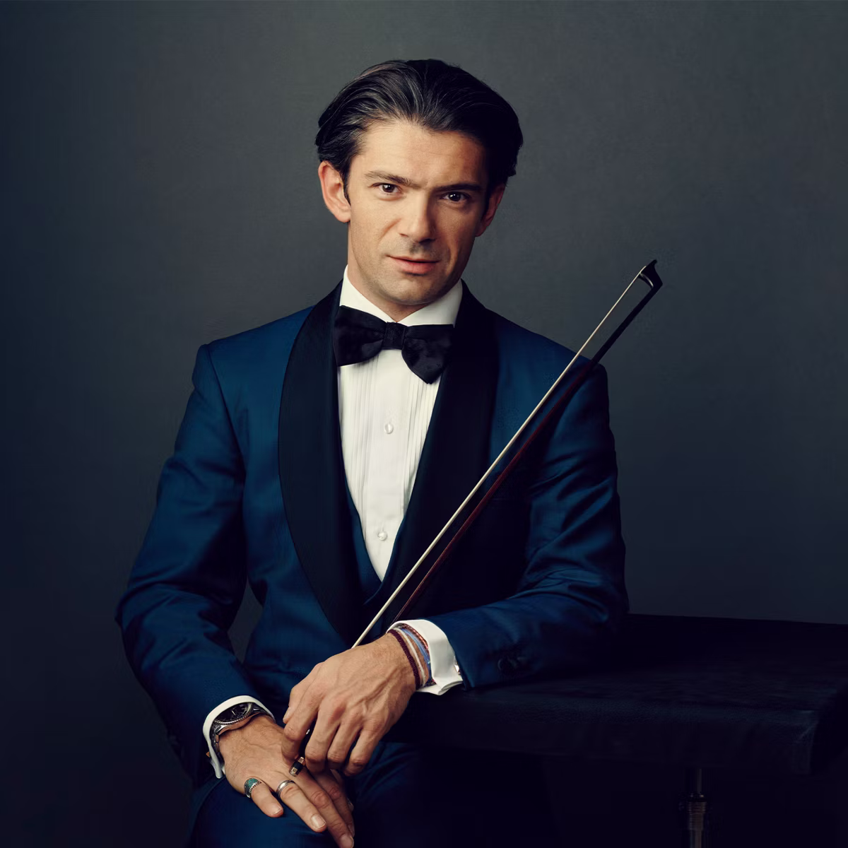 Smiling man in tuxedo holding an instrumental bow, posed against a dark studio background.