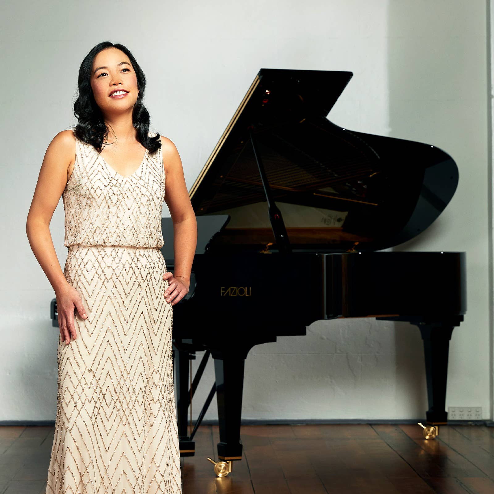 Woman in a beaded gown standing beside a black grand piano, smiling against a light wall background.
