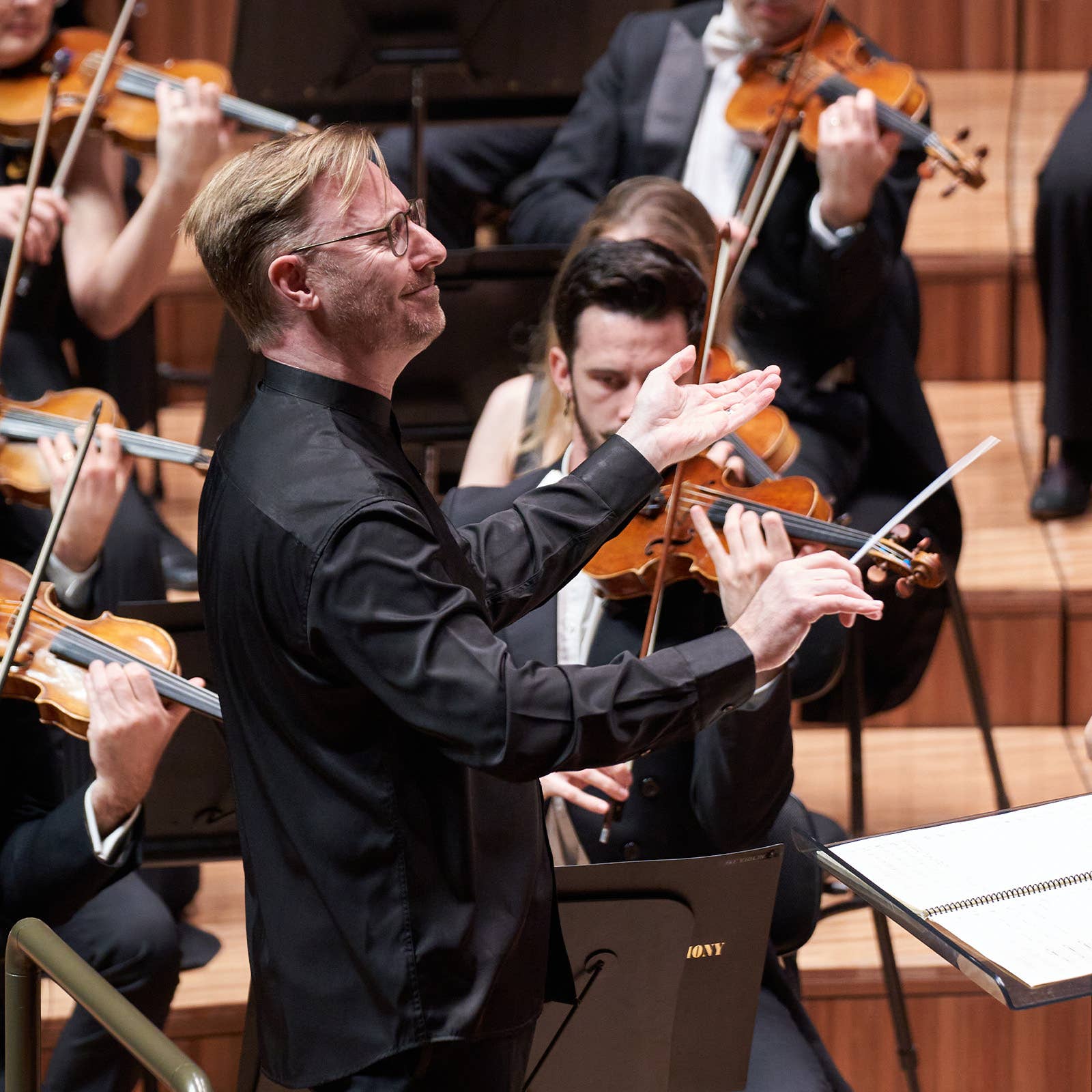Conductor in black gestures while violinists play in an orchestra on a wooden stage.
