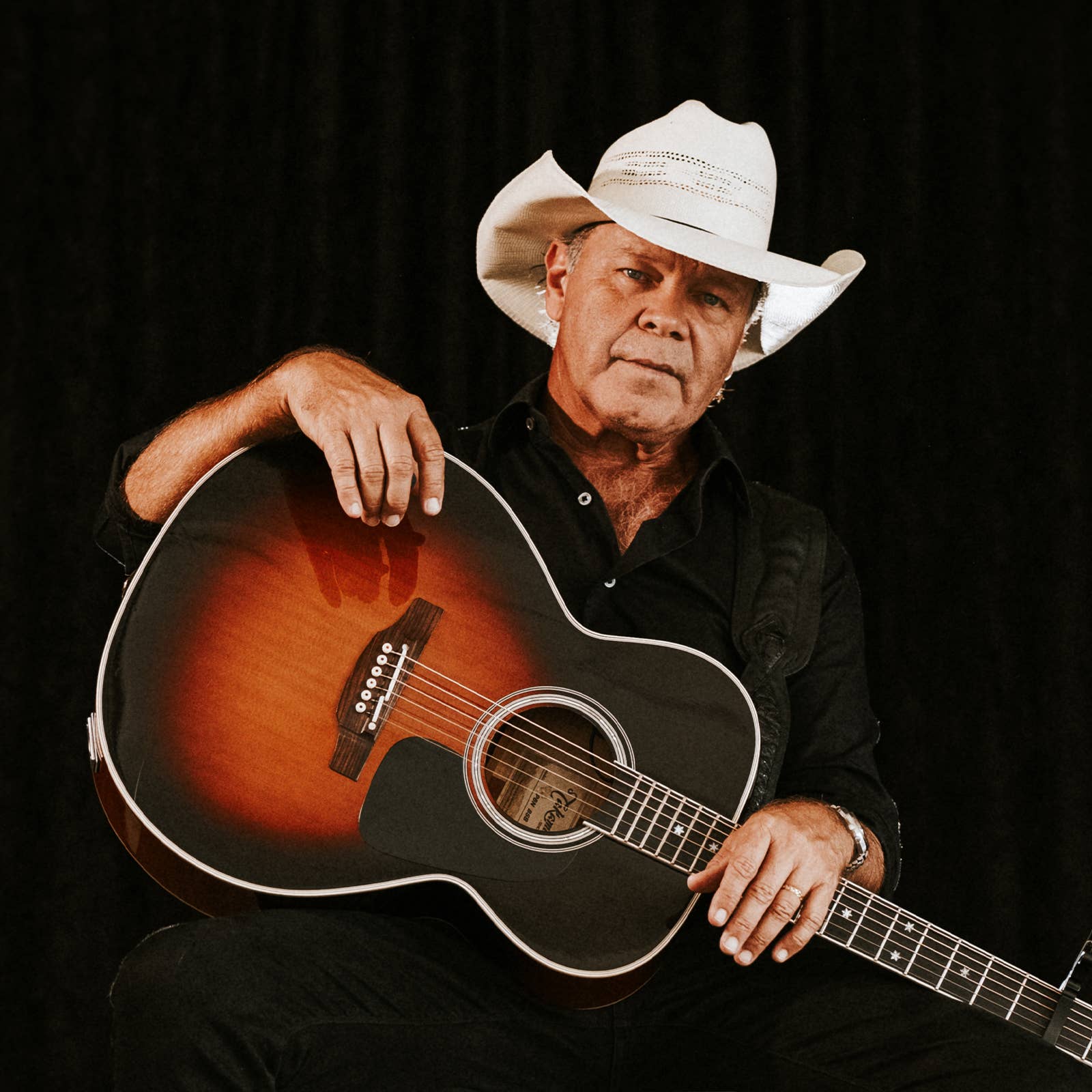 Older man in a white cowboy hat holding an acoustic guitar, hand on the strings against a dark background.