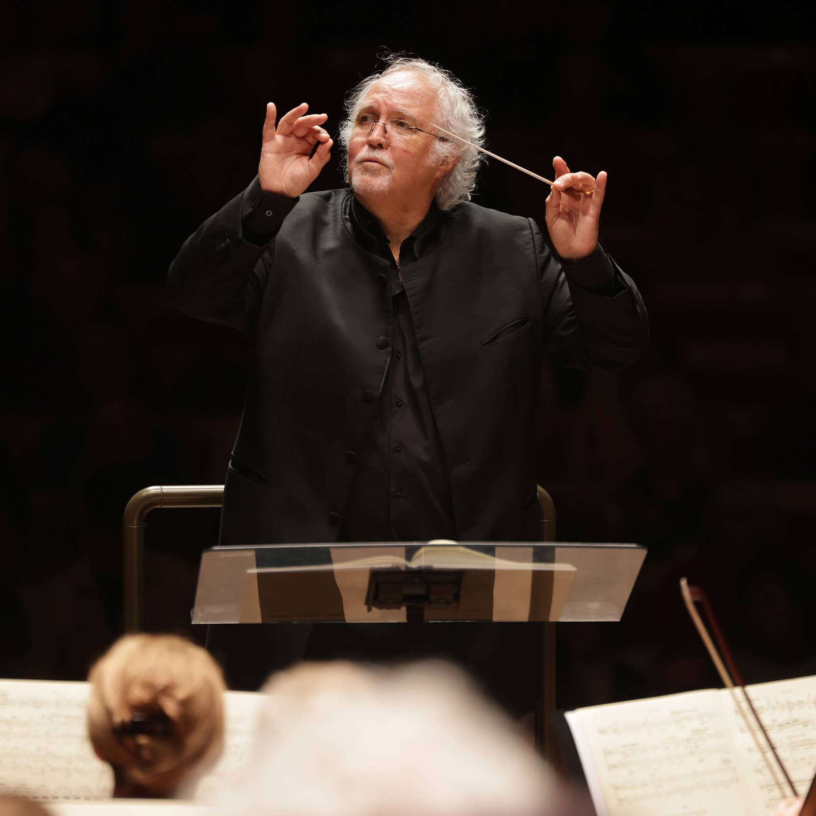 Conductor with white hair raising both hands, holding a baton during a live orchestra performance.