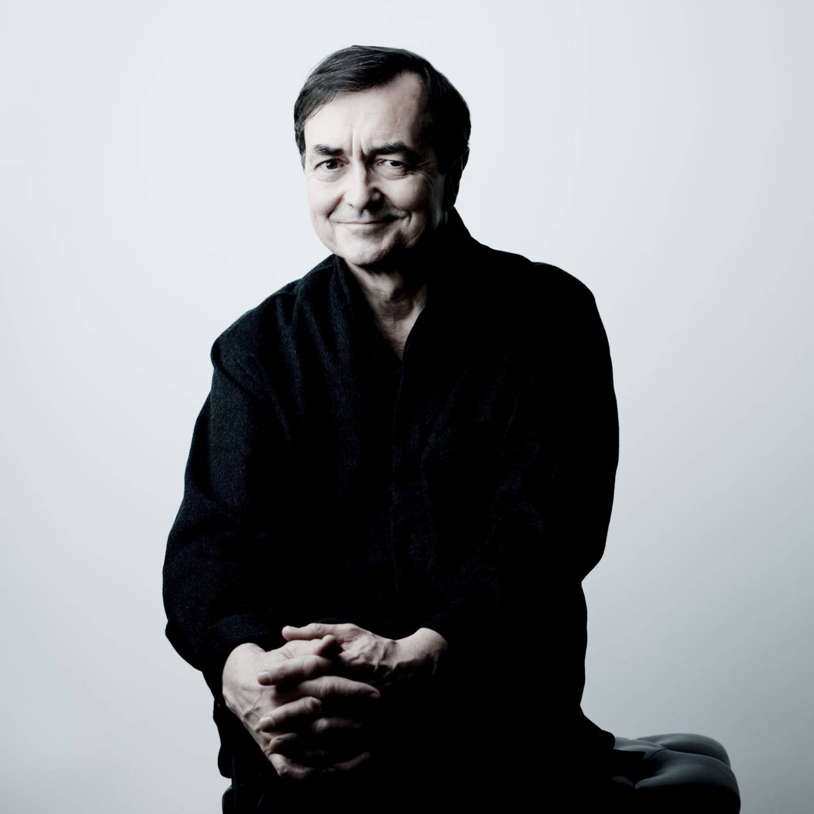 Smiling older man portrait, seated with hands clasped, dark sweater against a light gray background