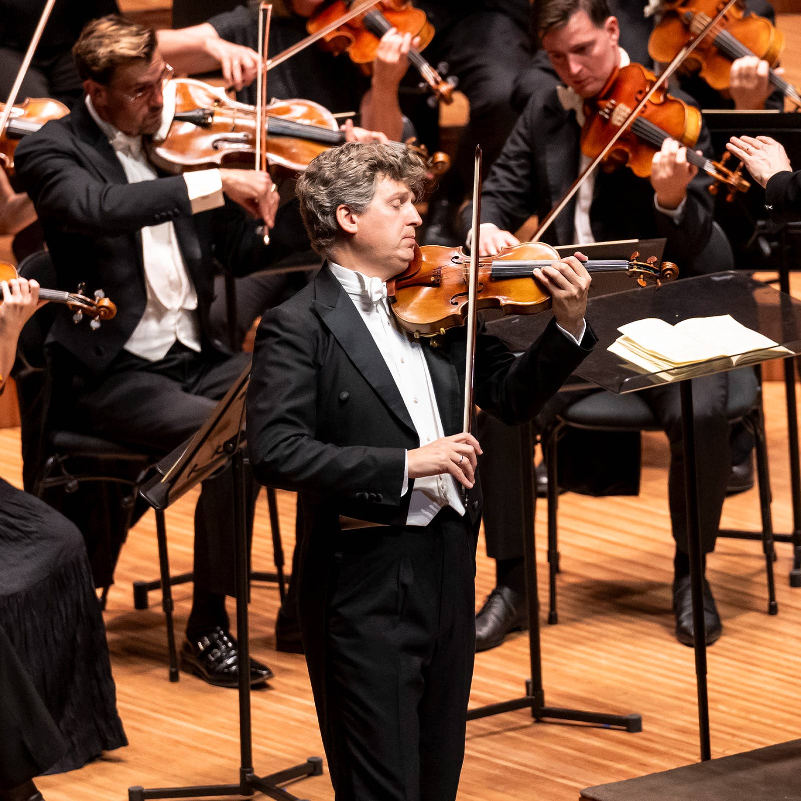 Violinist in black suit plays on stage with orchestra members behind him holding violins and sheet music.