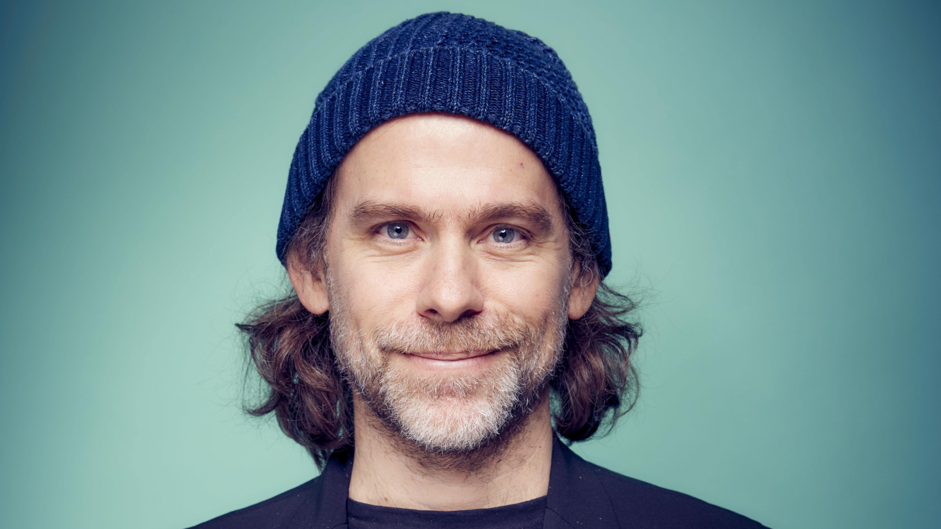 Man with gray beard wearing a blue knit beanie against a soft teal background.