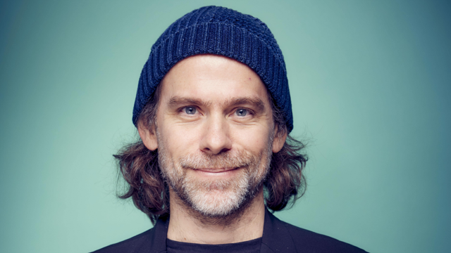 Man with gray beard wearing a blue knit beanie against a soft teal background.
