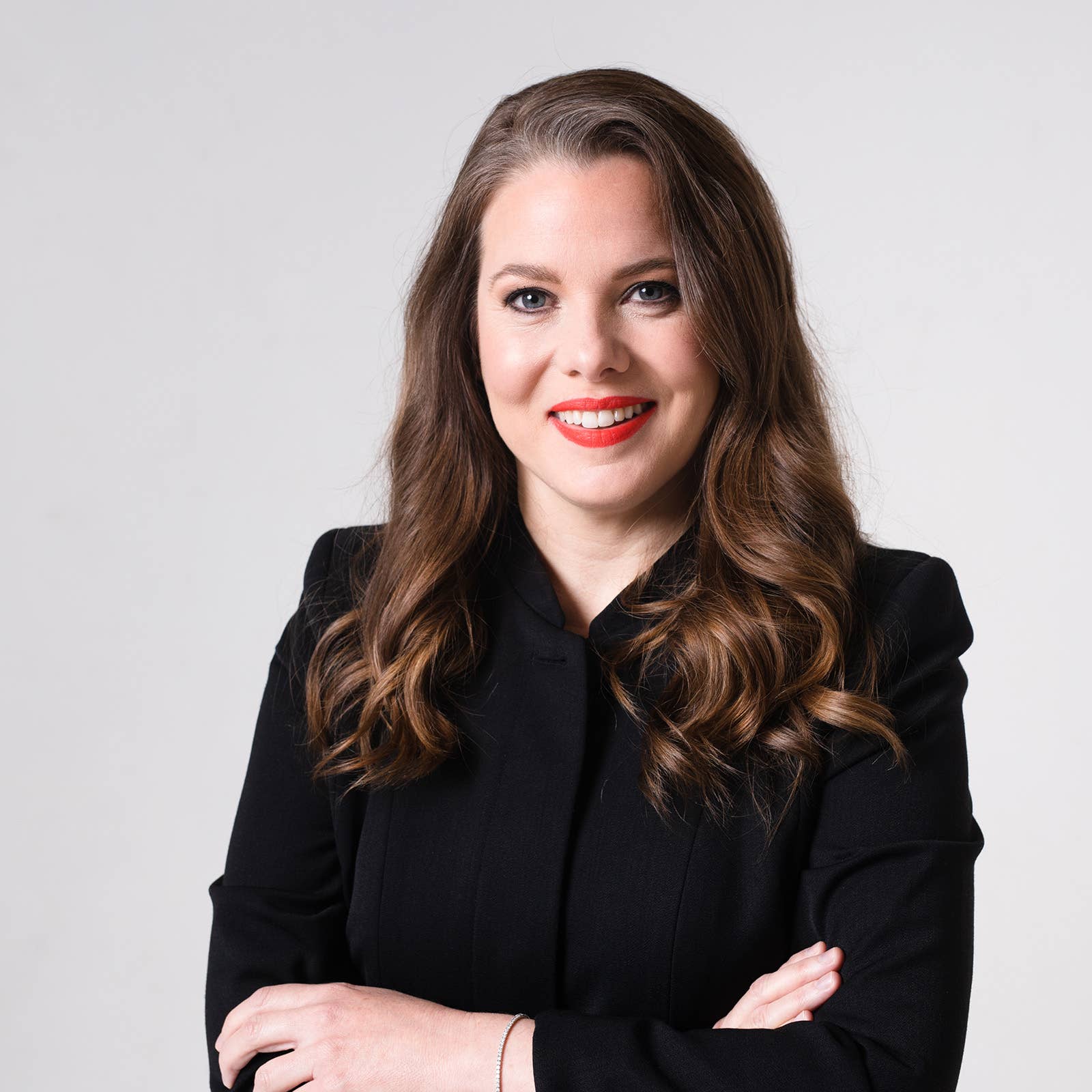 Woman in a black blazer with long brown hair and red lipstick smiling against a light gray background.