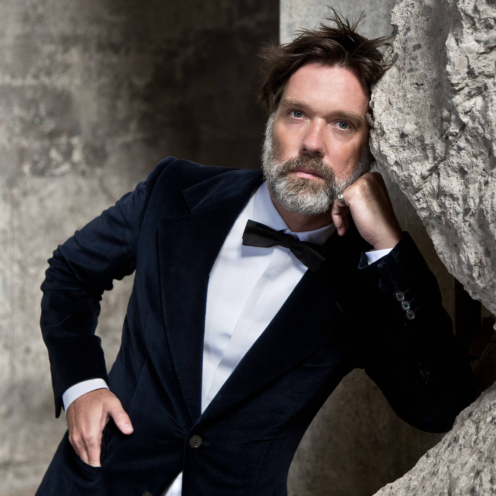 Bearded man in a tuxedo leaning against a textured stone, resting his head on his fist
