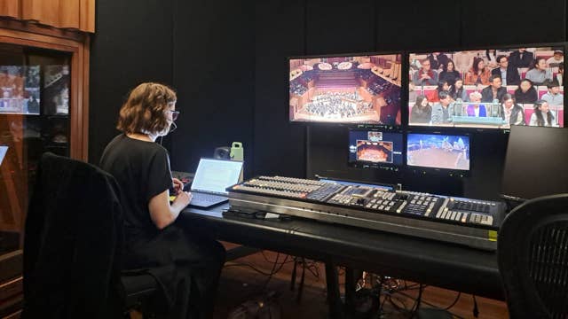Stage control room operator monitoring multiple screens with an audio mixing console and video feeds.