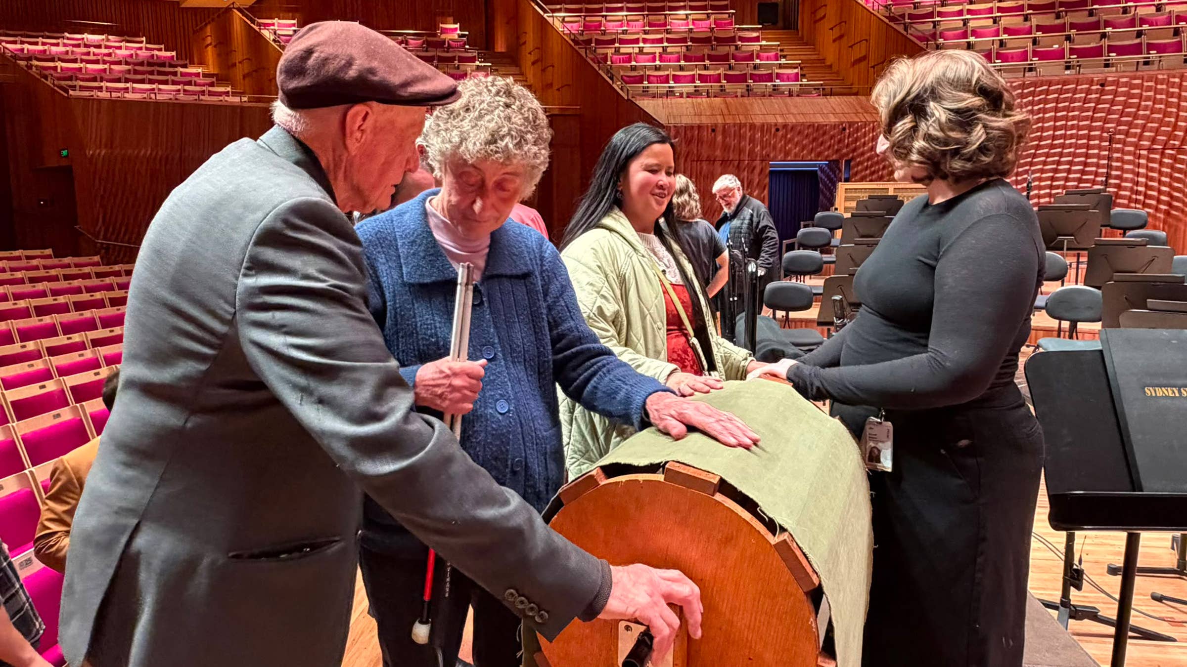 Elderly audience and volunteers in a theater, one woman touching a cylindrical instrument out while musicians sit nearby.