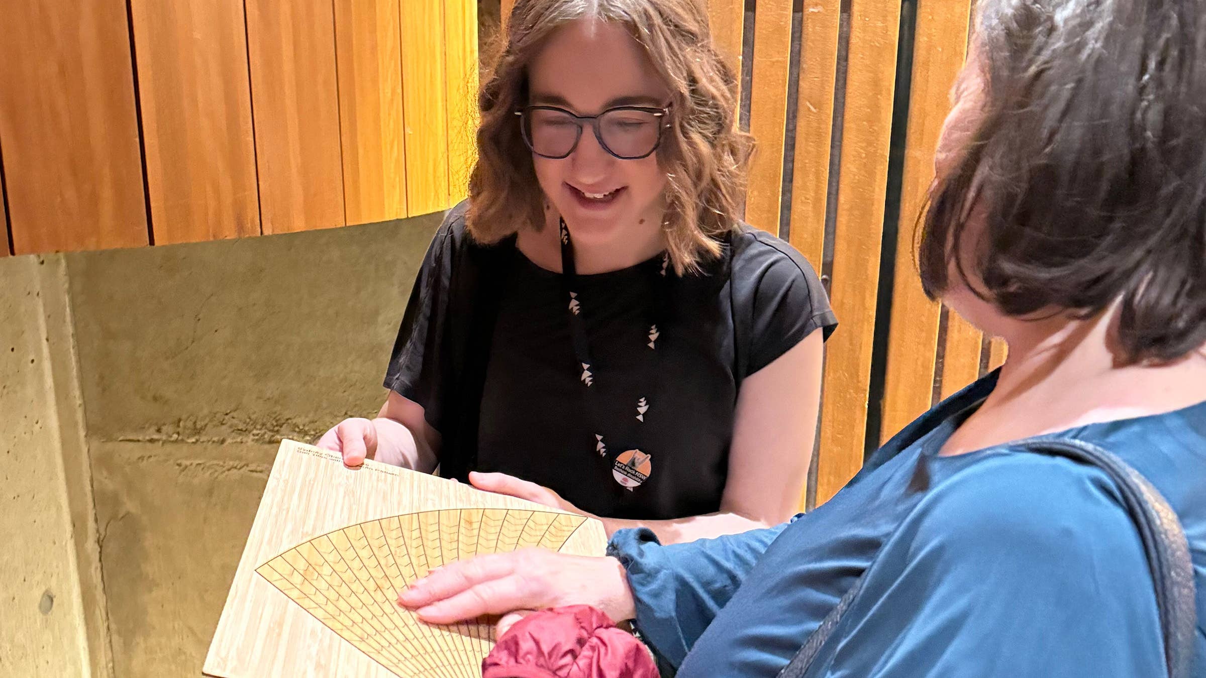 Smiling woman with glasses holds a cardboard fan while another person reaches in, outdoors by wooden panels.
