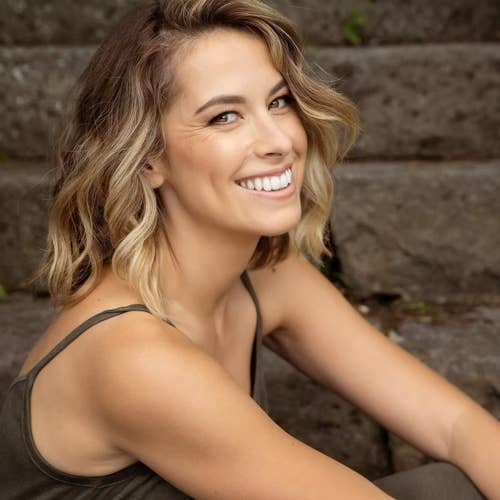 Smiling blonde woman in a dark tank top taking a selfie outdoors, with a brick wall background.