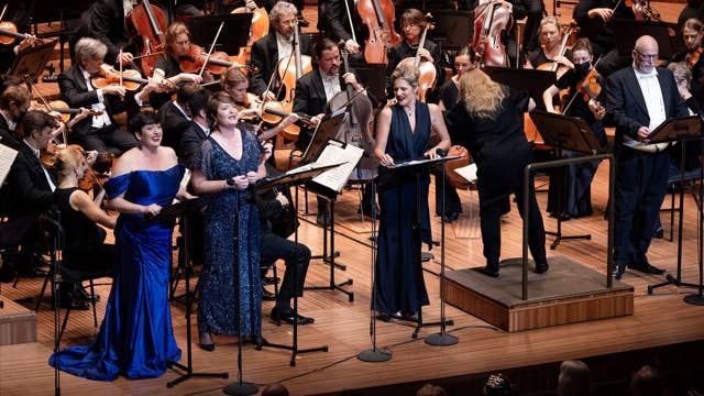 Orchestra stage with musicians playing violins and cellos beside three singers.