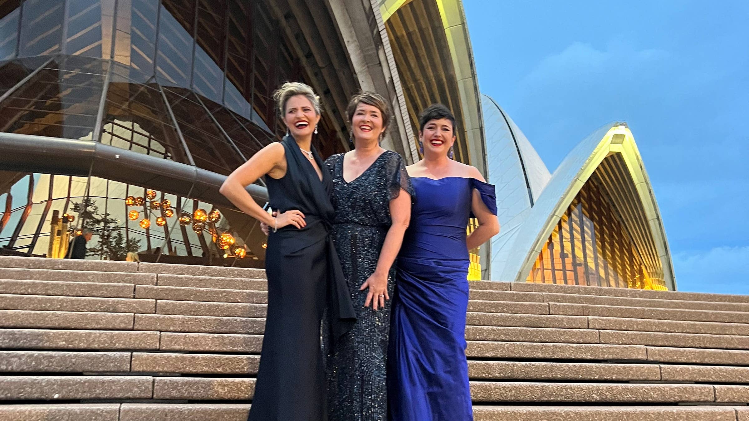 Three women in formal dark-blue gowns posing and smiling on Sydney Opera House steps under a clear blue sky