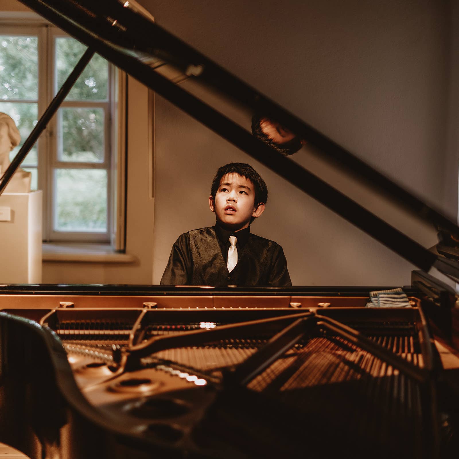 Young boy in formalwear sits at a piano, looking up under warm light by a window.