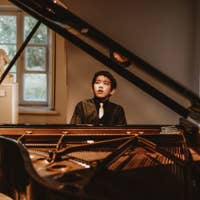 Young boy in formalwear sits at a piano, looking up under warm light by a window.