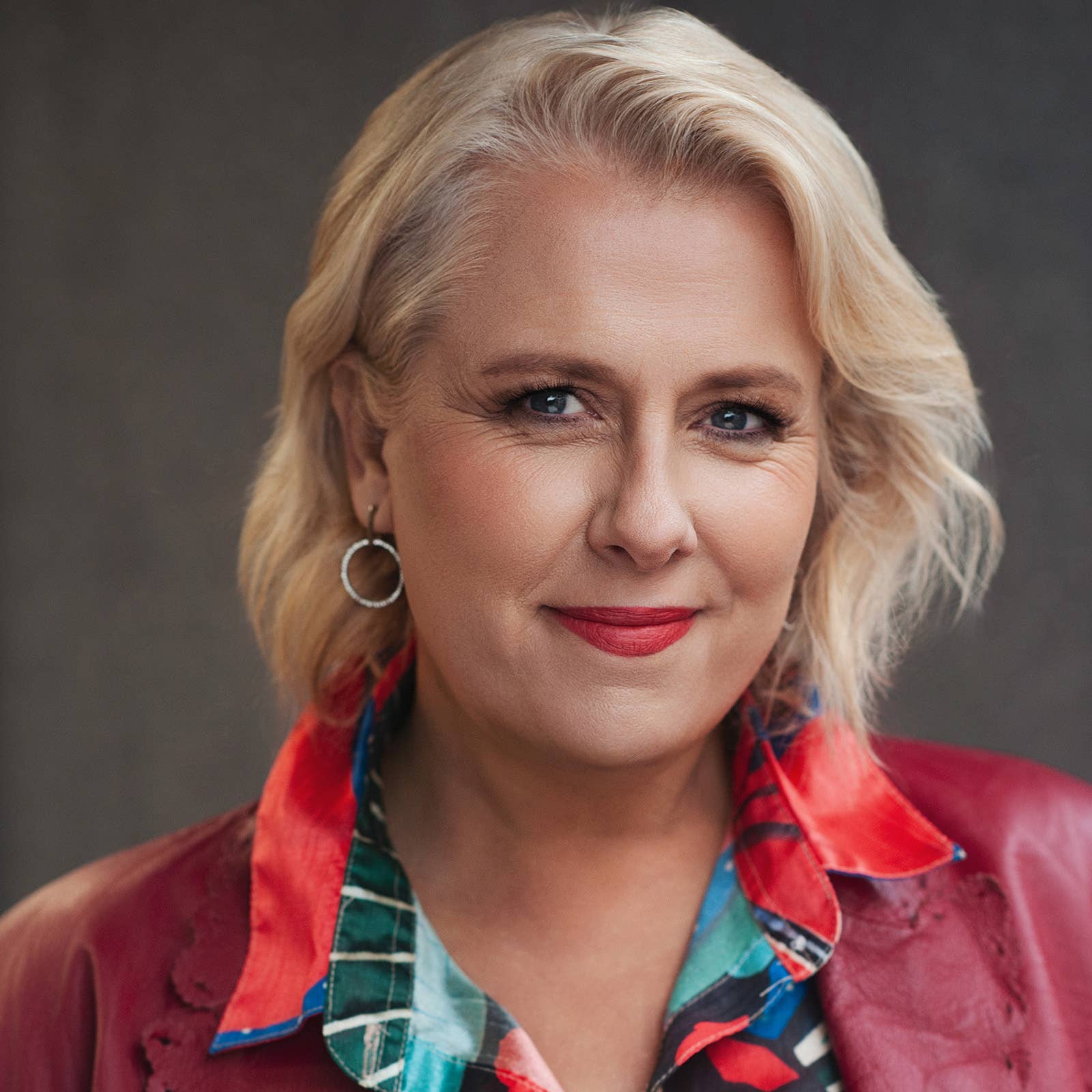 Close-up portrait of a smiling woman with blonde hair, wearing red lipstick and hoop earrings