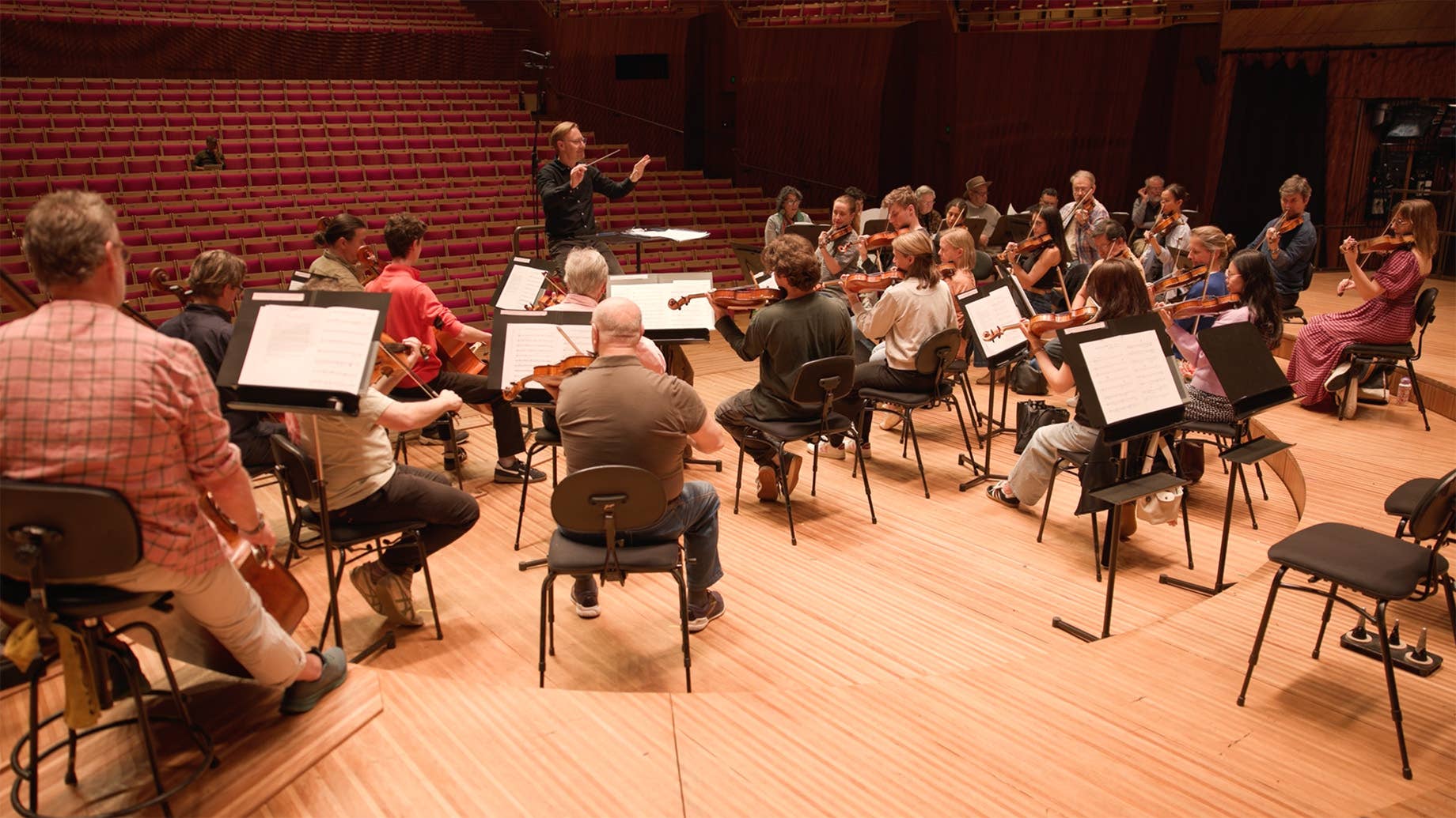 Orchestra seated on stage with conductor facing musicians in a red-seated concert hall, violins and cellos visible