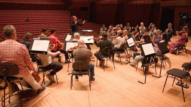 Orchestra seated on stage with conductor facing musicians in a red-seated concert hall, violins and cellos visible