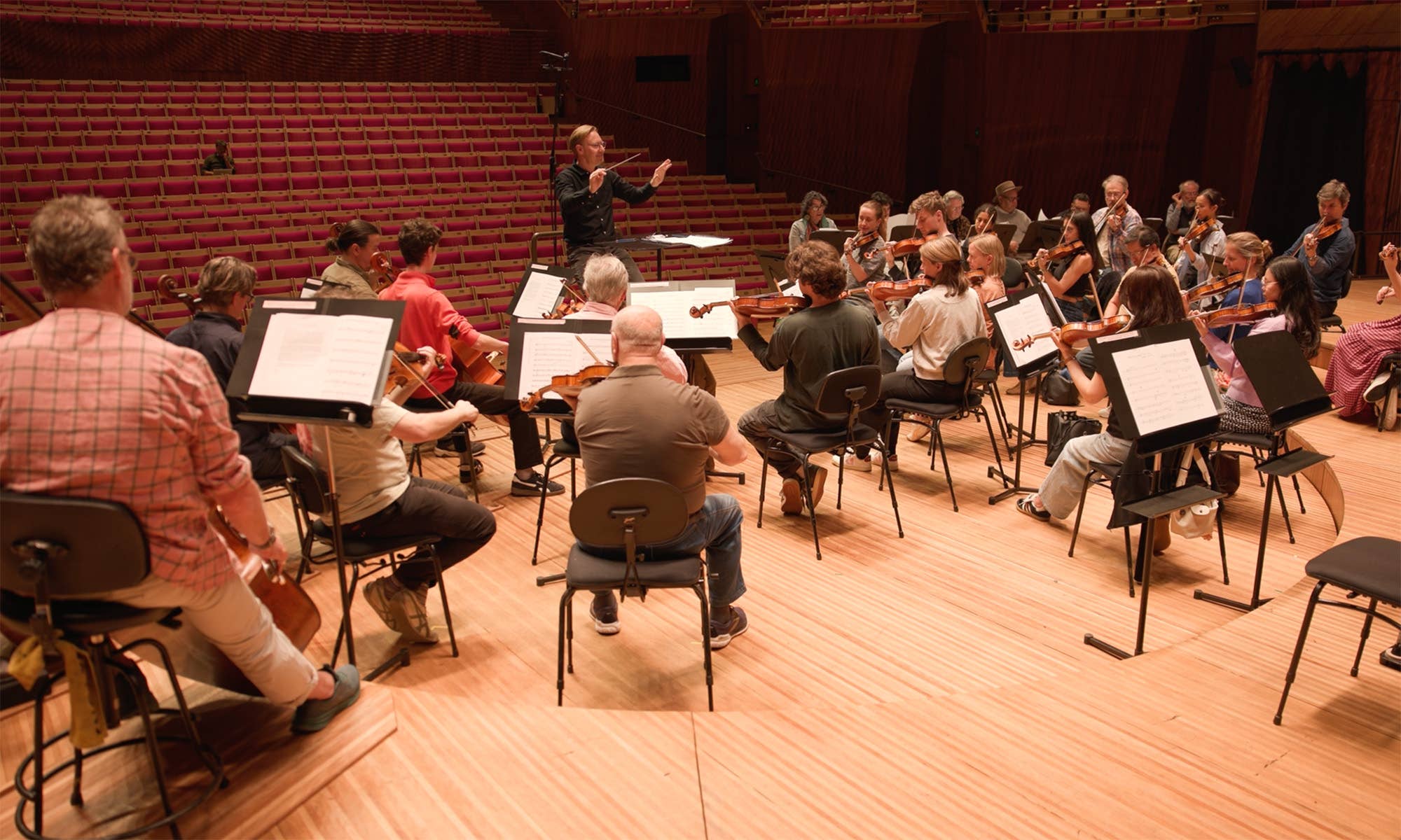Orchestra rehearsal in a concert hall with conductor leading string and wind musicians in red-seated auditorium