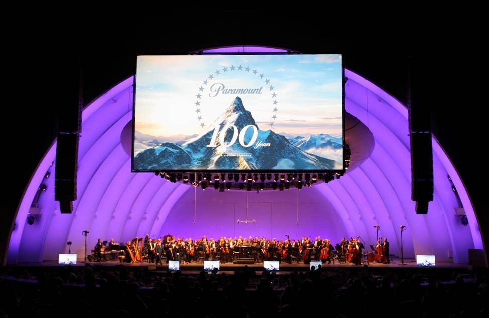 Concert stage with orchestra under purple lighting, large screen shows Paramount 100 years mountain backdrop