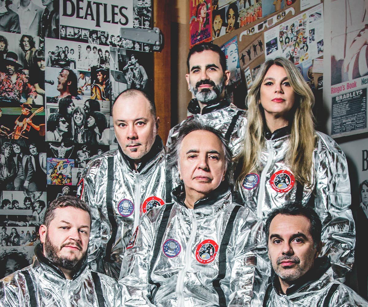 Members of Os Mutantes wearing aluminum spacesuits, behind them are posters of rock bands. 